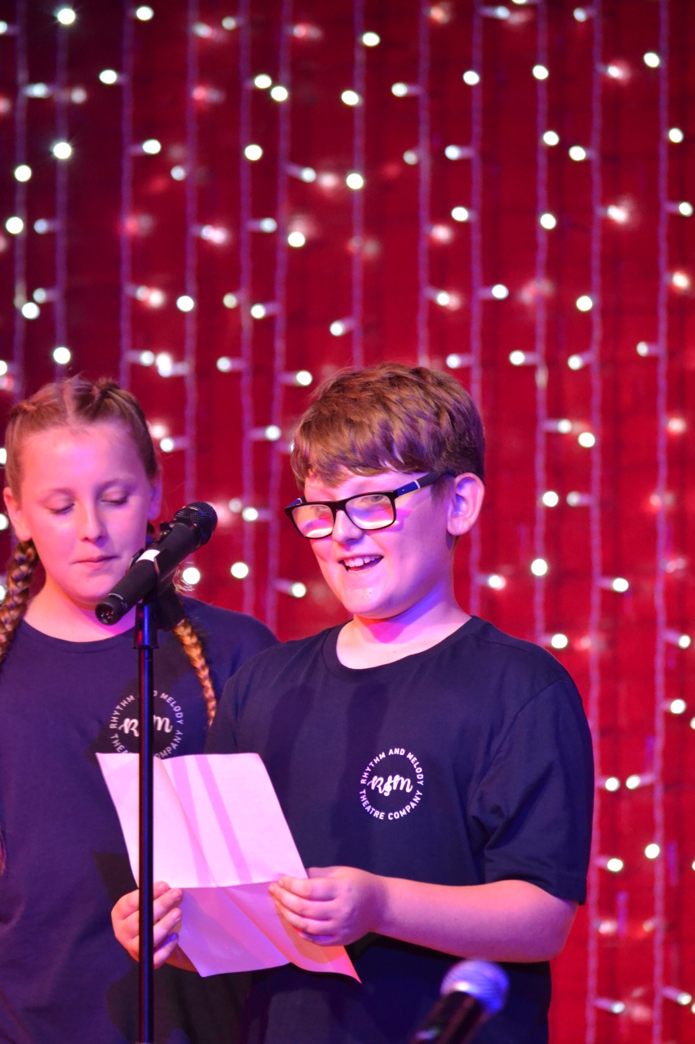 Two performers stand at a microphone, with one reading from a sheet of paper.