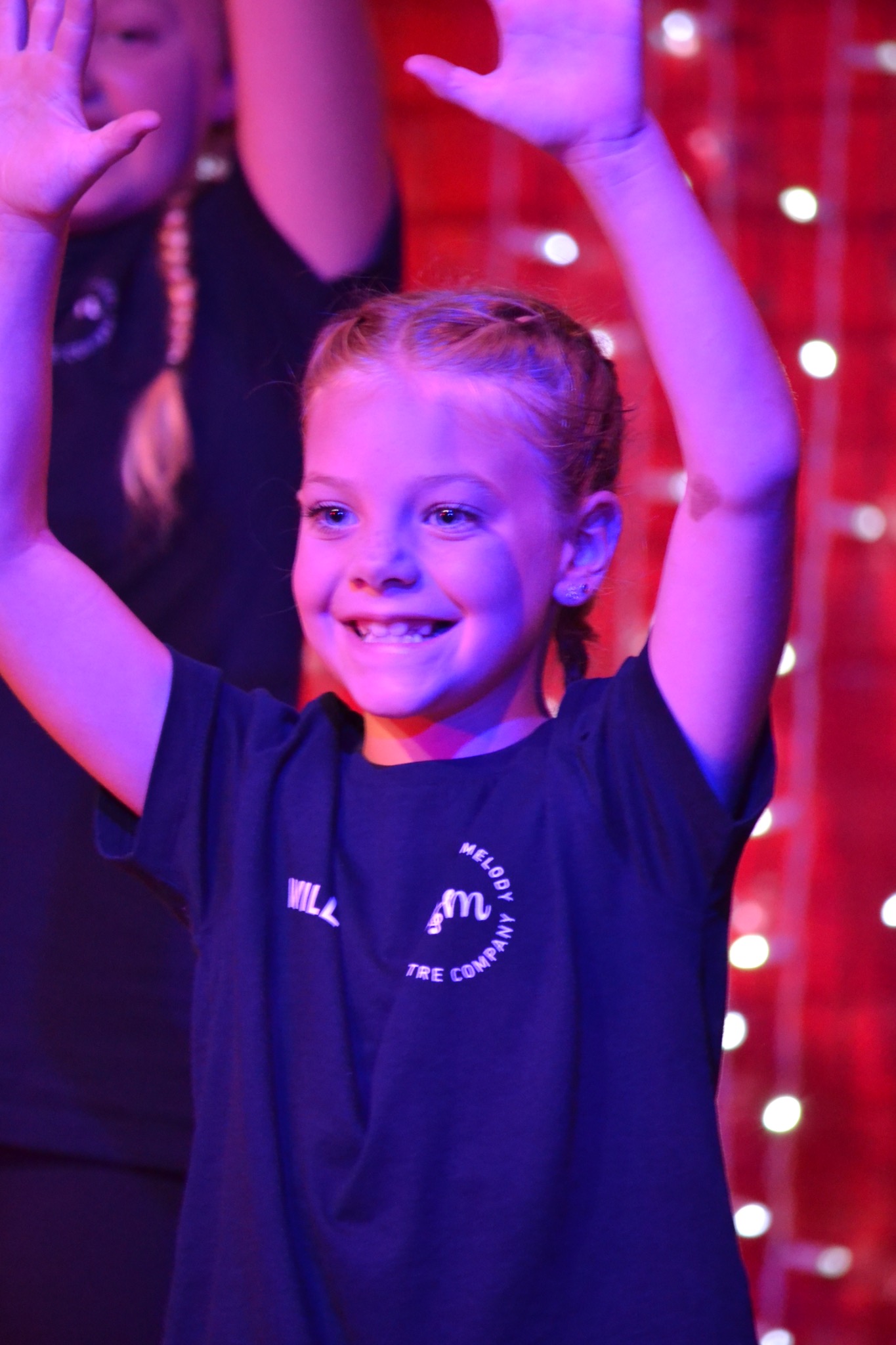 A smiling performer raises both hands beneath pink and purple stage lighting.